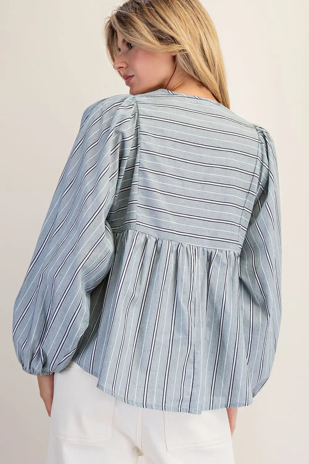 A woman shown from the back wearing a light blue and white vertically striped blouse with puffed balloon sleeves and a gathered waist seam, paired with white pants against a plain beige background.